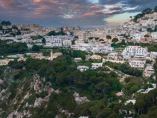 Capri Island in Italy features whitewashed buildings on a hillside, surrounded by greenery, rocky cliffs, and a dramatic orange and purple sky. © Aerial Film Studio