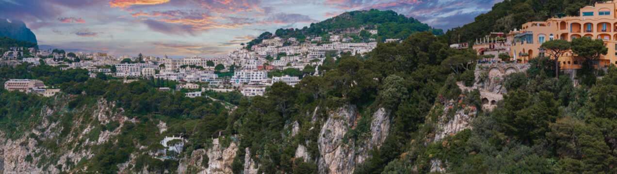 View of Capri town on Capri Island, Italy, with whitewashed buildings, lush greenery, rocky cliffs, and a prominent orange building at sunset.