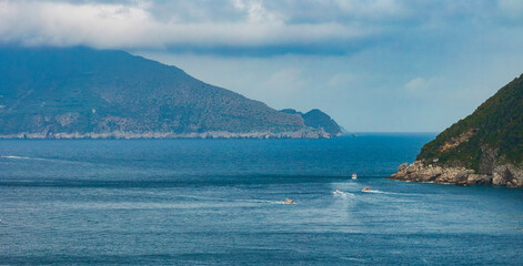 Fototapeta premium Serene coastal scene on Capri Island, Italy, with green cliffs, blue sea, small boats leaving trails, and a distant mountainous landmass under cloudy skies.