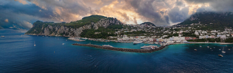 Capri Island's harbor features yachts, a curved breakwater, and clear waters. Whitewashed buildings sit on green hills below dramatic limestone cliffs. © Aerial Film Studio