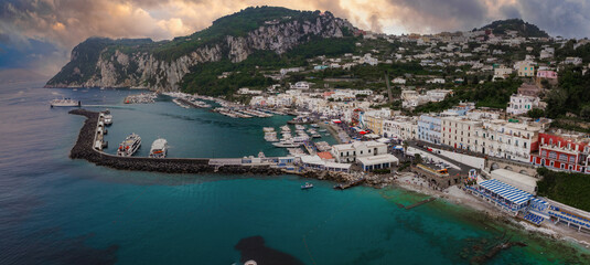 Turquoise waters, boats, and ferries at Marina Grande on Capri Island, Italy. Whitewashed buildings and greenery cover the hillside under an overcast sky. © Aerial Film Studio