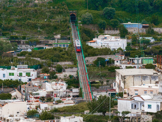 A red funicular car ascends a steep track on Capri Island, Italy, surrounded by lush greenery,...