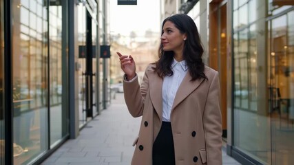 A young adult woman with dark hair, dressed in a white shirt and beige trench coat, strolls through a modern city arcade. She smiles and points, engaging with a presumed augmented reality overlay. - Powered by Adobe
