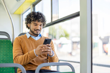 A young, smiling man uses his phone while riding public transport in a bright setting.