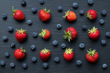 Strawberries and blueberries lie on a gray slate. The fruits are photographed from above. The strawberries glow red.