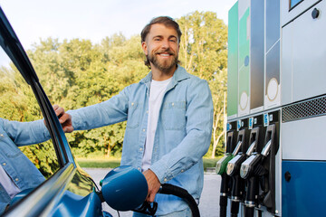 Happy handsome man filling up his automobile with fuel, standing near is car at modern petrol station, smiling at camera