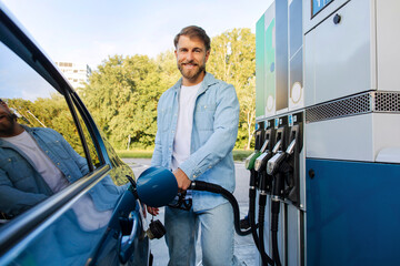 Happy European man refueling car at gas station, standing near automobile and holding fuel nozzle,...