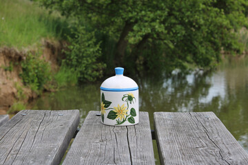 A painted jar with a lid on the edge of a wooden pier by the water at the shore of a pond.