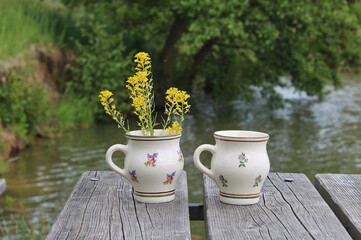 Two painted ceramic country mugs on the boards of a wooden pier by the pond, one of which contains...