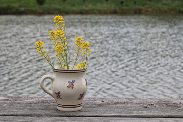 Painted ceramic mug with a plant, the yellow herb Barbarea vulgaris, by the water at the edge of...