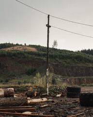 Abandoned Power Pole with Lines in the Raw Quarry Landscape with a Forest in the Background under the Cloudy Sky.