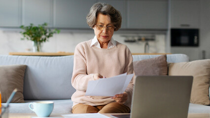 Focused senior woman sitting at table, using laptop, banking online or checking utility bills,...