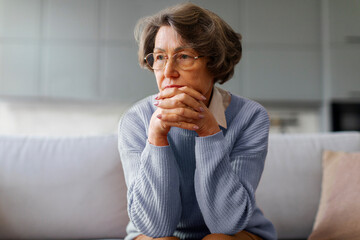 Depressed senior lady touching chin, sitting on sofa and looking away, feeling worried and...