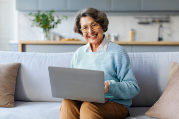 Happy European retired woman using laptop online for remote shopping, sitting on couch at home and smiling at camera. Grandmother working freelancing