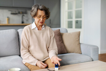 Senior woman with pulse oximeter on hand measuring oxygen saturation level, sitting on couch at...