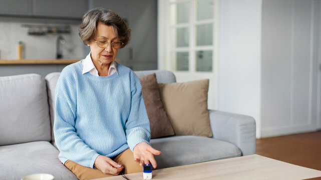 Senior woman with pulse oximeter on hand measuring oxygen saturation level, sitting on couch at home. Pulse oxymeter medical device, healthcare on retirement concept