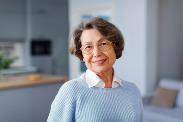 Pleasant elderly European woman looking at camera with warm smile, posing for photo in modern home interior. Ageless beauty concept