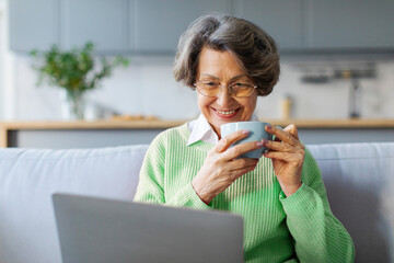 Positive gray-haired grandma drinking coffee and watching favorite show on laptop or video chatting...