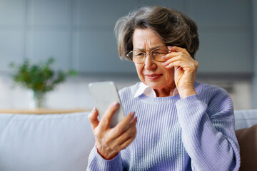 Frustrated senior retired woman in glasses getting problems with smartphone, trouble with online connection, reading bad news, sitting on couch