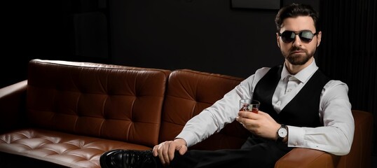 Handsome man in formal clothes drinking whiskey while sitting on sofa in dark room