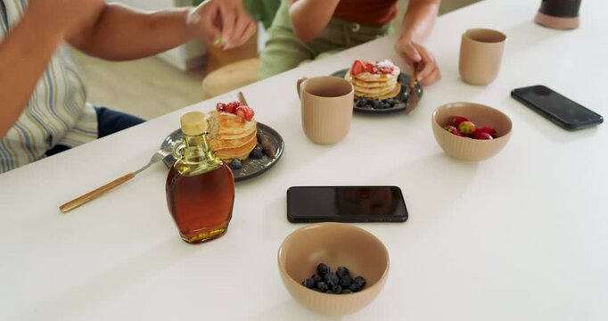 Diverse couple enjoying breakfast with pancakes and berries at home, sharing meal