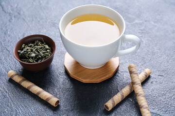 Drink coasters with cup of tea, dry leaves in bowl and wafer rolls on dark table