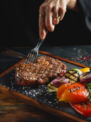 Woman eating grilled ribeye steak served on a dark slate plate with vegetables, scene with cutlery - fork and knife