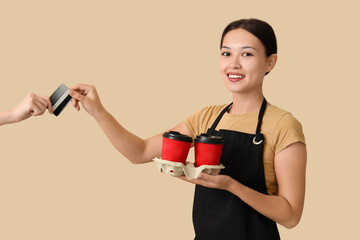 Female Asian barista with cups of coffee taking credit card on beige background