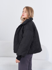 Female model in a black puffer jacket with a high collar, adjusting the collar with her right hand....