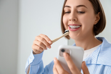 Young woman with dental braces and mobile phone brushing teeth in bathroom, closeup