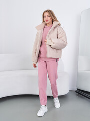 Young woman in pink tracksuit with matching puffer jacket stands by a mirror. She wears white...