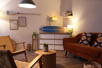 Interior of stylish living room with brown sofa, armchairs, glowing lamps and surfboard at evening