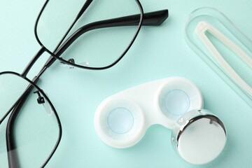 Contact lenses in case, tweezers and glasses on light turquoise background, flat lay