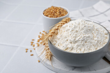 Wheat flour, grain and spikes on white tiled table, closeup. Space for text