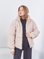 A woman wearing a beige puffer jacket over a dark hoodie stands against a plain setting. She gazes...