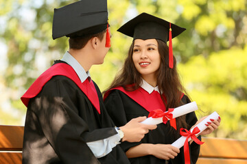 Couple of graduating students with diplomas sitting on bench outdoors