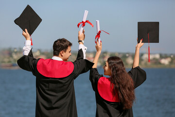 Couple of happy graduating students with diplomas on embankment, back view
