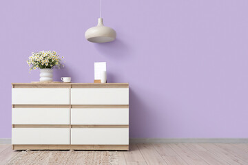 Vase with chamomiles and coffee cup on chest of drawers near lilac wall in room