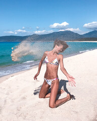Portrait of beautiful woman wearing bikini, while sand flying against her face on the beach. Sporty...