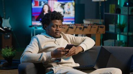 Relaxed African American freelancer smiling while browsing his smartphone on a comfortable couch....