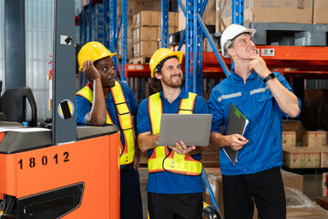 Three male warehouse workers from diverse ethnicities standing beside forklift, smiling and looking upward with thoughtful expressions, wearing safety gear while holding laptop and clipboard.