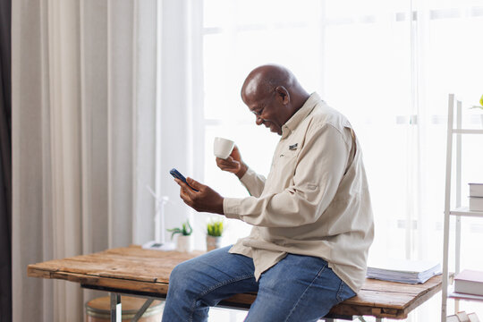 Portrait of smiling mature man using mobile phone, watching video and holding cup of coffee, sitting at workplace. Guy browsing internet, surfing web, free copy space