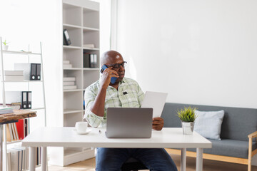 Busy mature middle aged business man, senior professional executive manager talking on the phone making business call on cellphone sitting at work desk in office using laptop computer.
