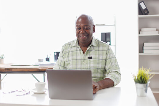 Senior businessman sitting alone in his home office and working on laptop computer. Smiling black man using laptop in living room. Businessman using phone laptop at home.
