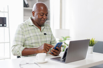 Senior businessman sitting alone in his home office and working on laptop computer. Smiling black man using laptop in living room. Businessman using phone laptop at home.
