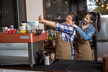 Young Asian female barista massaging shoulders of senior Asian woman barista in brown aprons inside...
