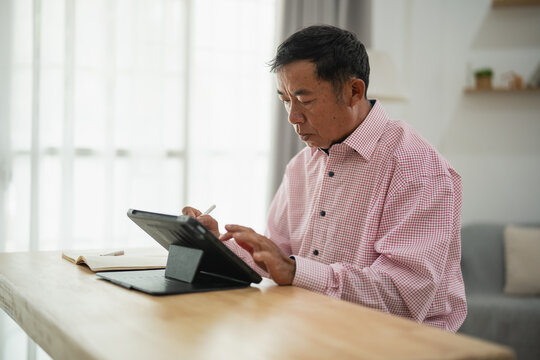 Focused senior man wearing checkered shirt using tablet while sitting at modern wooden table in bright room with natural light and cozy atmosphere