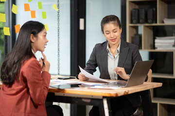 Two Asian businesswomen holding graph papers are sitting and talking happily while working.