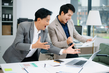 Asian businessmen in modern office working on reports and data using laptop, tablet, and phone, shaking hands, celebrating success, finance and teamwork concept.

