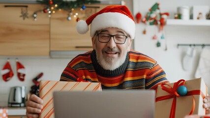 This festive image captures a man with a Santa hat and glasses, smiling as he holds gifts during a video call on a laptop in his decorated kitchen, spreading holiday cheer. - Powered by Adobe